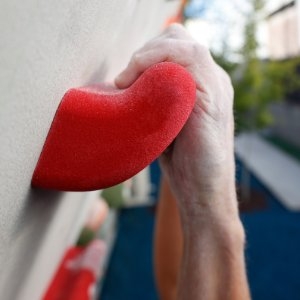 A hand gripping a large red climbing hold mounted on an outdoor climbing wall. The hold has a rounded, ergonomic shape.